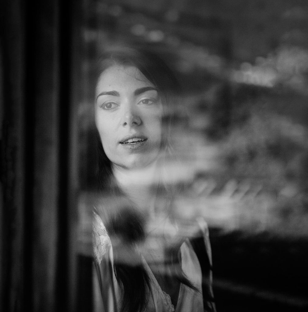 Ritratto in bianco e nero di una donna che guarda fuori da una finestra, con il suo riflesso che si sovrappone al vetro creando un’atmosfera intima e contemplativa.