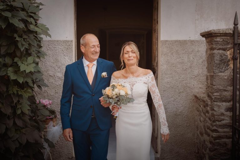 La sposa esce di casa con il padre, trattenendo le lacrime per l’emozione del momento, mentre lui le sorride orgoglioso prima di accompagnarla alla cerimonia.