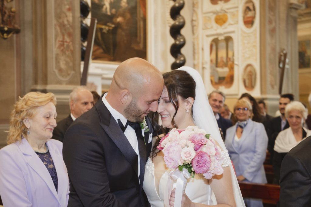 Gli sposi si abbracciano in chiesa con le fronti unite, emozionati e sorridenti, mentre gli invitati li osservano commossi durante la cerimonia.