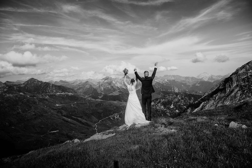 Gli sposi festeggiano sulla cima della montagna con le braccia alzate, osservando il panorama vasto delle vette alpine sotto un cielo nuvoloso.