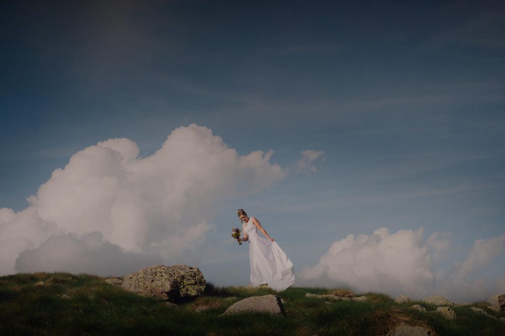 La sposa posa su un’altura erbosa tra rocce e nuvole imponenti, illuminata da una luce morbida di montagna.