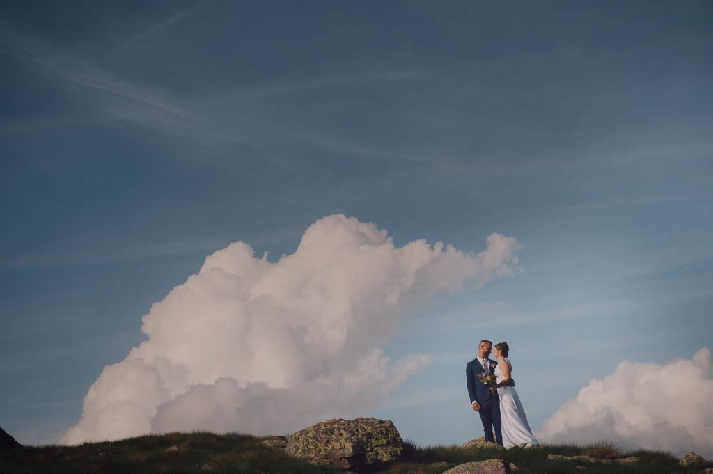 Gli sposi si guardano negli occhi in cima alla montagna, con grandi nuvole bianche che riempiono il cielo alle loro spalle.