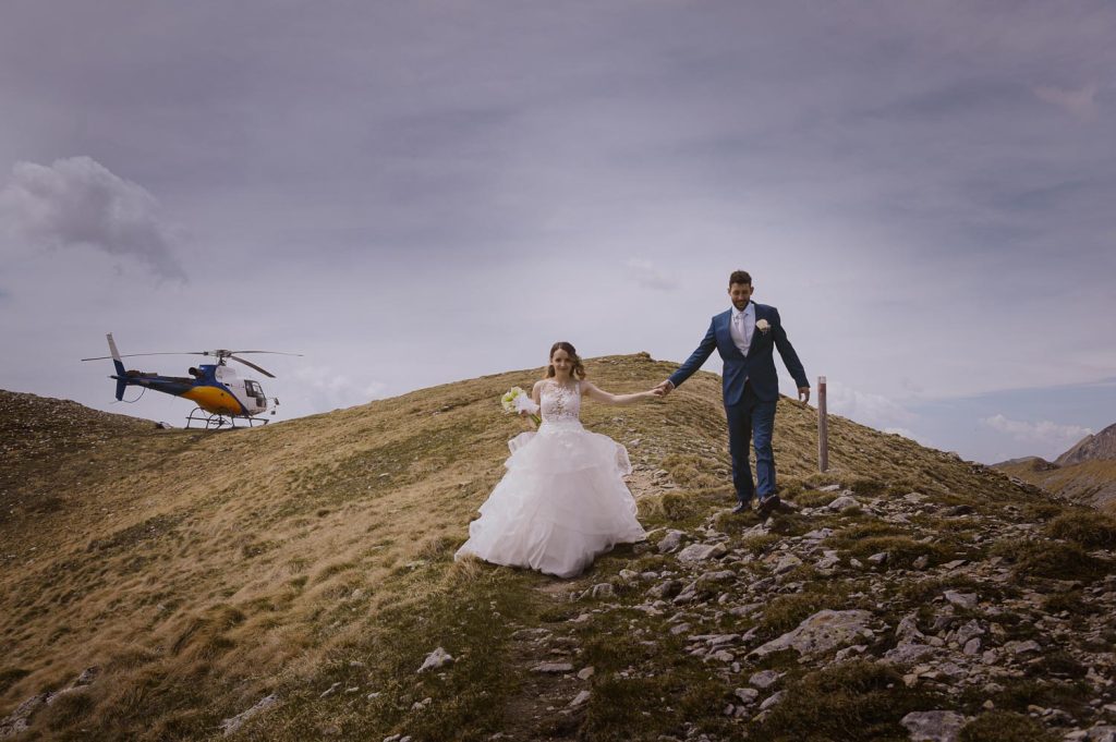 Gli sposi camminano mano nella mano su un crinale di montagna, con un elicottero posato sullo sfondo, durante un servizio fotografico in alta quota.