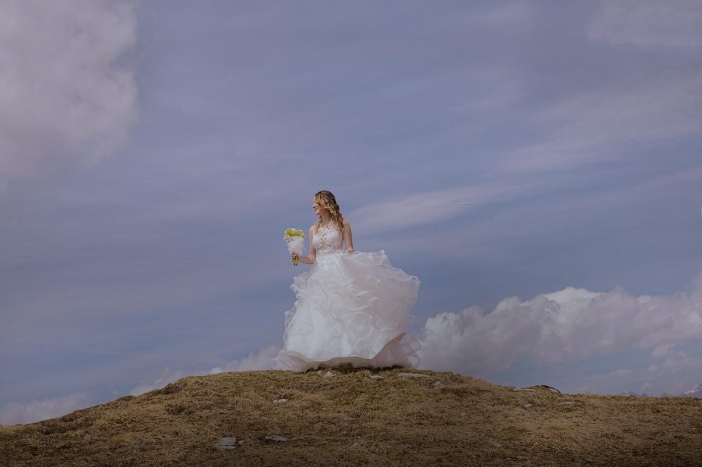 La sposa posa sola sulla cima della montagna, guardando il bouquet mentre il panorama ampio del lago e delle catene montuose si apre alle sue spalle.
