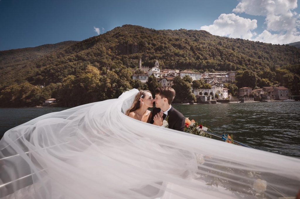 Sposi che si baciano in barca avvolti dal lungo velo con il borgo di Corenno Plinio affacciato sul Lago di Como.
